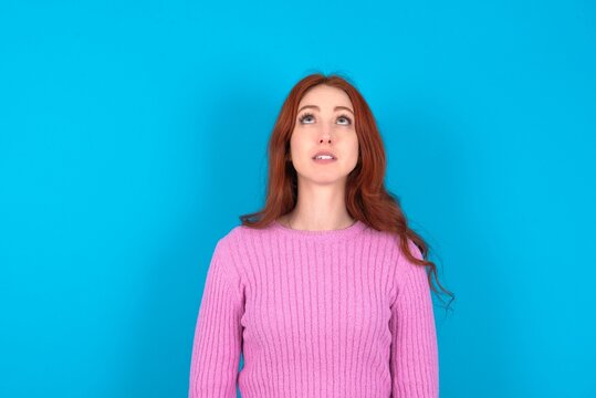 young woman wearing pink sweater over blue background looking up as he sees something strange.
