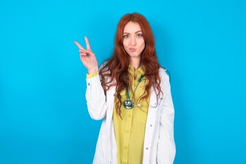 young doctor woman wearing medical uniform over blue background  makes peace gesture keeps lips folded shows v sign. Body language concept