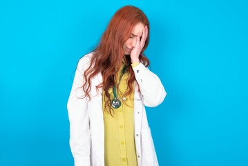 young doctor woman wearing medical uniform over blue background with sad expression covering face with hands while crying. Depression concept.