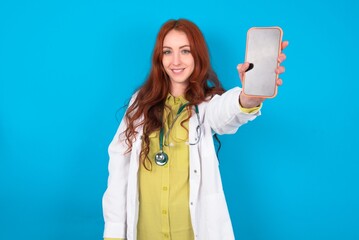 Charming adorable young doctor woman wearing medical uniform over blue background holding modern device, showing black screen smartphone