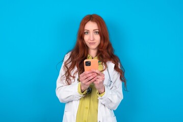 Portrait of serious confident young doctor woman wearing medical uniform over blue background holding phone in two hands