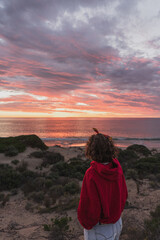 Fototapeta premium Women with short hair and red hoodie stand up in the sand closed to the beach and looking to the sea and sunset