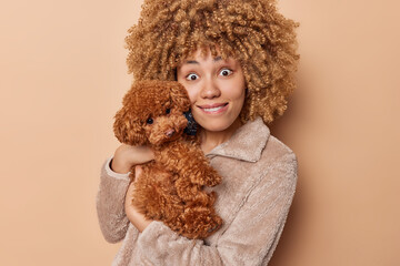 Portrait of surprised pleasant looking woman with bushy curly hair bites lips poses with small pedigree poodle puppy chills with favorite pet dressed in winter outerwear isolated over beige background