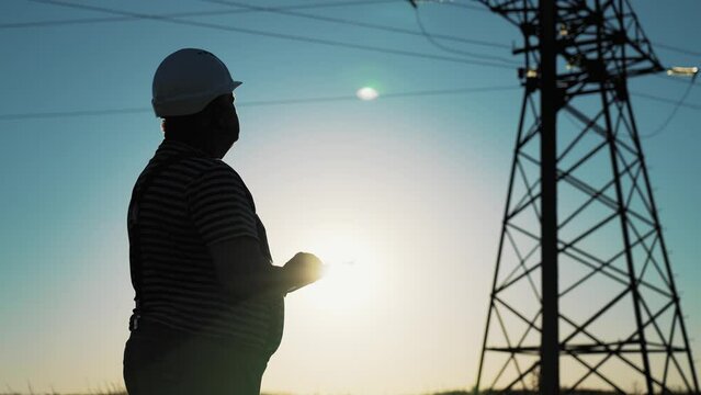 Silhouette businessman man in protective helmet in field using tablet and looks at telecommunications tower. Construction worker stands against blue sky. Online work with technology for connection.