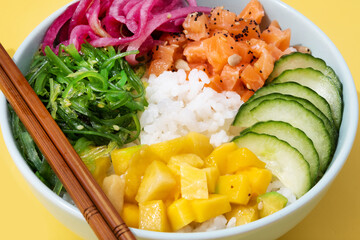 Poke bowl with rice, salmon,cucumber,mango,onion,wakame salad, poppy seeds ands sunflowers seeds on yellow background. Close up