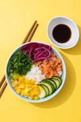 Poke bowl with rice, salmon,cucumber,mango,onion,wakame salad, poppy seeds ands sunflowers seeds on yellow background