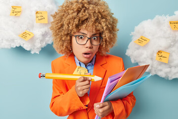 Beautiful shocked businesswoman holds big pencil and folders does different tasks wears spectacles...