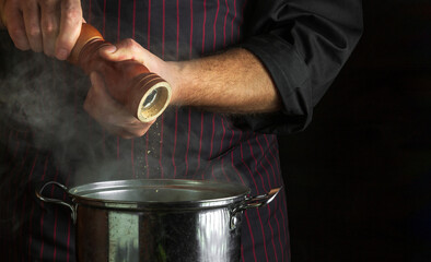 Professional chef adds ground pepper to a pot of boiling food. Retsoran kitchen cooking concept with advertising space on dark background