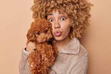 Friendship and domestic pets care. Surprised curly haired woman pouts lips holds poodle puppy near...