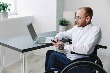 A man wheelchair in the office works at a laptop with a phone, working online, social networks and startup, integration into society, the concept working person with disabilities, real person close-up