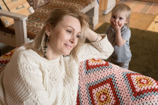 Adult Blond Woman Sit On Sofa At Home Relaxing While Small Daughter Play Alone