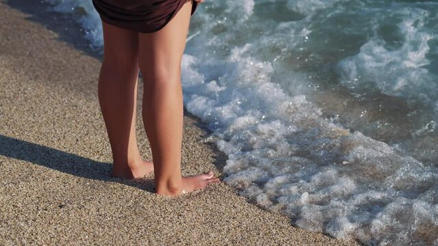 Legs feet of young female walking sand barefoot on the beach. Slim female legs and feet walking along sea water waves on sandy beach. Pretty woman walks at seaside surf. Summer vacation in ocean.