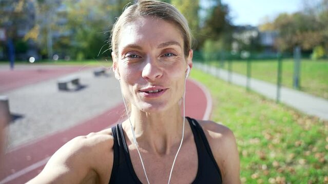 POV Close up. Sportswoman talking on a video call using a smartphone while standing in a stadium. Woman in headphones looks at the camera and shares positive impressions about training with her friend