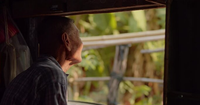 An Asian male farmer who is a lonely elderly employee working in a rural farm sitting and looking through the old narrow window and his face lit up in the morning light.