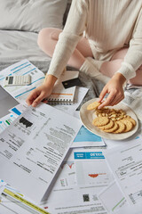 Unrecognizable woman manages expenses and finances studies bills and documents dressed in domestic clothes eats cookies works from home. Unknown person surrounded by cheque bills and papers invoices