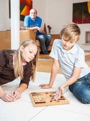 Family Life, Sibling Rivalry. A brother and sister engrossed in a game of backgammon at home while father watches. From a series of related images.