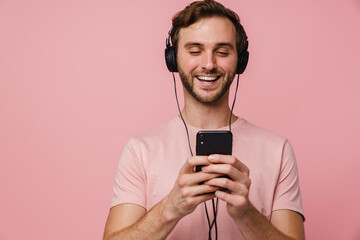 Smiling young man listening music with headphones and mobile phone isolated over pink wall