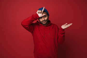 Indian man doing loser gesture and holding copyspace on palm isolated over red background