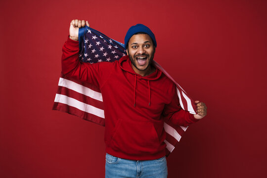 Cheerful indian man holding US flag while standing isolated over red background