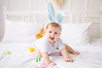 cute yawning baby boy with bunny ears and colorful eggs on a white bed at home playing, little...