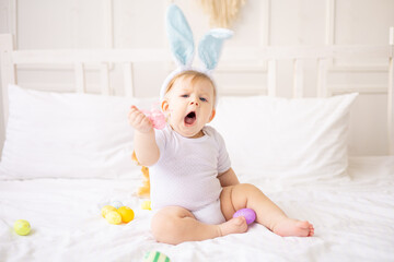 Fototapeta premium cute yawning baby boy with bunny ears and colorful eggs on a white bed at home playing, little blonde baby with a rabbit, happy easter concept