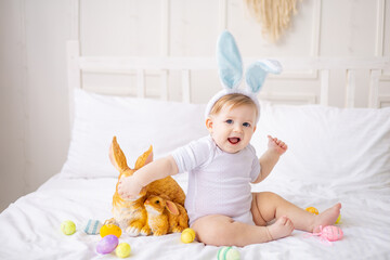 cute baby boy with bunny ears and colorful eggs on a white bed at home playing, little blonde baby with a rabbit, happy easter concept - Powered by Adobe