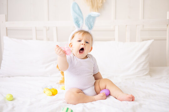 cute yawning baby boy with bunny ears and colorful eggs on a white bed at home playing, little blonde baby with a rabbit, happy easter concept