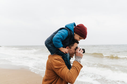Smiling father carrying on shoulders his son and taking pictures while spending time together on beach