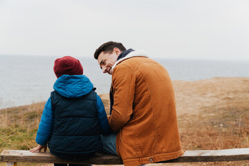 Back view of father and his son spending time together while sitting on bench at beach