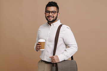 Young indian guy smiling while standing with cup of coffee and bag isolated over beige wall
