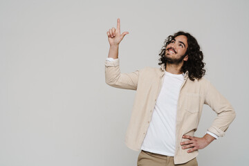 Smiling hispanic man pointing finger up at copy space while standing isolated over white wall