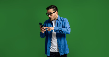 Surprised man in denim shirt using smartphone on green background