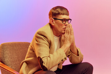 Portrait of mature, middle-aged man in jacket sitting on chair with thoughtful face, posing over gradient pink purple background in neon light. Concept of emotions, lifestyle, facial expression, fun