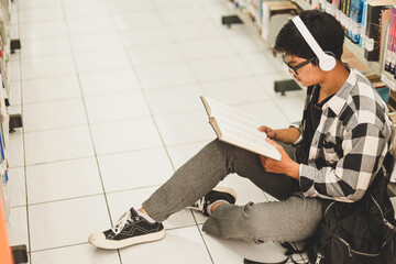 Young student in casual style wearing backpack is sitting on the floor, reading a book by bookshelf while listening music on headset in the library 