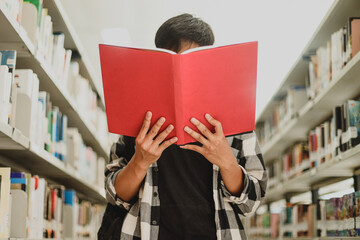 Portrait of a young student in casual style reading a book and covering his face by bookshelf in...