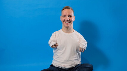 a smiling and cheerful man in a white T-shirt with disabilities sits on an isolated blue background.