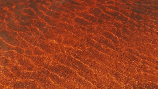 Transparent red water as abstract background, top view. Ripples on the surface of the water from the wind or breeze. Sandy light bottom. Surreal horrifying background. Blood or claret