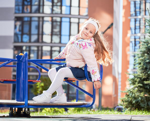 Fototapeta premium Little female child whirling on merry-go-round, having fun at playground. Front view of adorable girl on blue swing, smiling, while revolving outdoors. Modern residential buildings on background.