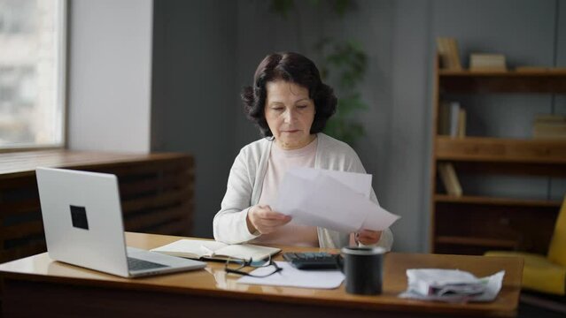 An elderly woman sits at a desk at home and studies payments or taxes. She checks the numbers in the documents and enters the data into the laptop. The concept of bill payments and tax calculation.