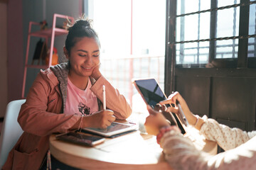 A young woman is drawing a design in her digital tablet while taking care of her sister in a coffee place