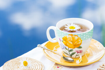 Herbal tea in teacup decorated with flowers outdoors in background of blue sky with clouds, healthy herbal chamomile drink with honey, tea drinking concept