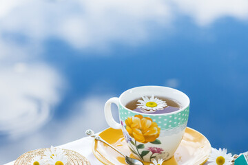 Herbal tea in teacup decorated with flowers outdoors in background of blue sky with clouds, healthy...