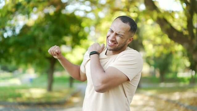 A young athletic man feels sharp pain in his shoulder during morning exercise in a city park. A sad man in a T-shirt was injured. The unfortunate athlete rubs and massages the muscles on the sore spot