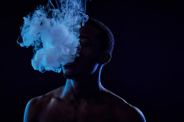 Young African American man standing in front of camera in darkness and blowing cloud of vapor out...