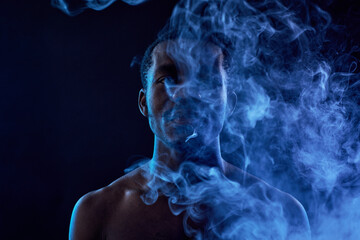 Young African American man hiding behind large cloud of smoke while standing in front of camera and releasing vapor out of his mouth