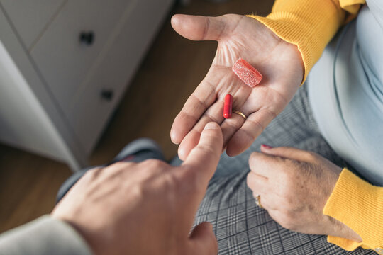 Palm image of an unrecognizable older woman in a yellow sweater sitting on her bed holding a red pill and a jelly bean being helped by another person to choose what to take as medication.