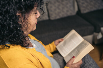 Side image of an older woman in a yellow sweater sitting on the sofa concentrating on reading a...
