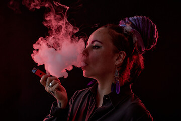 Young woman in casualwear holding electronic cigarette and blowing smoke out of her mouth while...