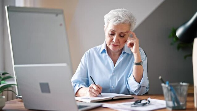 Mature adult european woman at office using laptop computer for work. Beautiful serious professional senior lady. Businesswoman sitting at office table at workplace. Boss office worker. Freelance.