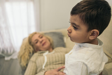 Obraz na płótnie Canvas Middle-aged blonde woman lying on the couch smiling and looking intently at her young autistic son sitting on her. Selective focus on the child.